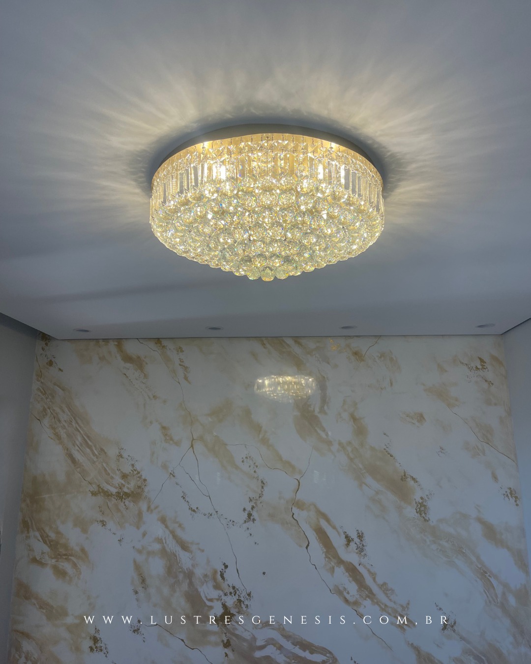 Lustre de cristal redondo Harion Ø50 Dourado com pedras transparente da Lustres Gênesis instalado no teto, iluminando ambiente sofisticado com parede marmorizada.